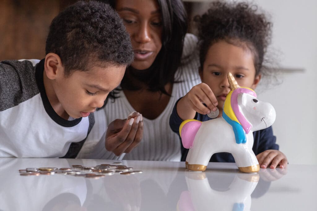 A beautiful mother of African American descent is teaching her two children of elementary school age the importance of saving money for a brighter future. The adorable children are depositing all of their pocket money to the unicorn shape money box.