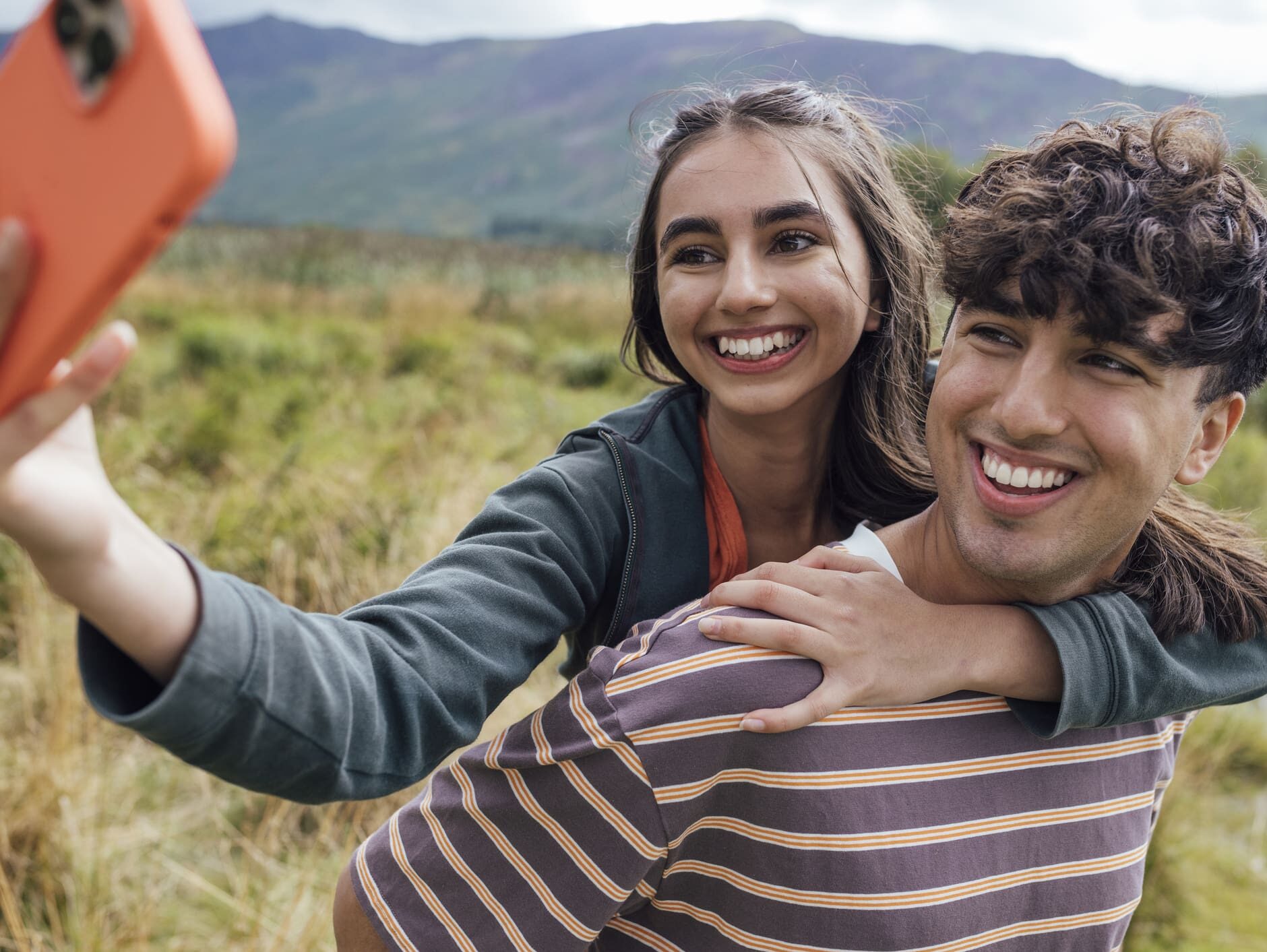 Brother and sister on a walk together through the countryside in Keswick, The Lakes. They are using a smart phone to take a selfie while they piggyback one another.