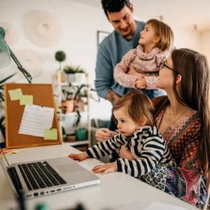 Working mom on laptop with baby on lap, while stay-at-home dad holds toddler nearby